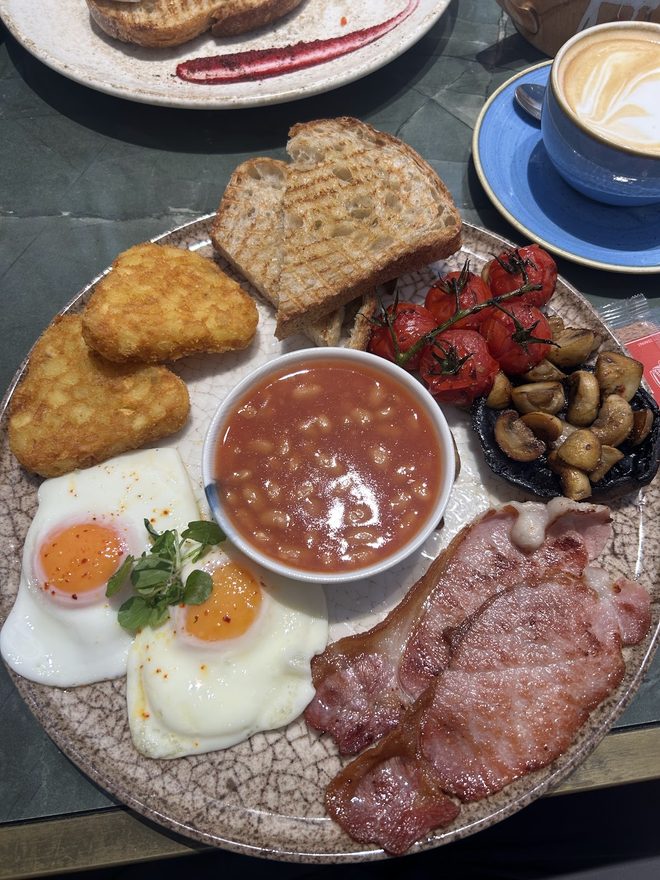 A cooked breakfast with fried eggs, bacon, hash browns and beans, cappuccino behind
